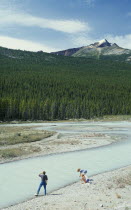 Visitors beside river looking towards dense pine forest with jagged mountain peak above.