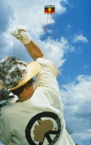 Person flying kite seen from behind at low angle looking up into blue sky with white clouds.