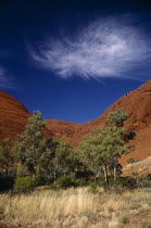 Also known as Katatjuta  meaning Many Heads. Large red rocks with trees in front and whispy white clouds in the sky above
