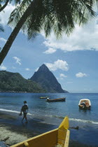 Man walking along beach with boats moored offshore and the Pitons behind.  Prow of boat and palm in foreground.Windward IslandsWindward Islands