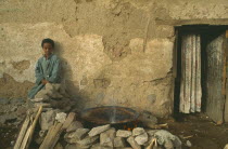 Child sitting outside poor household with injera cooking on open fire at side.