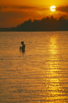 Line fisherman standing waist deep in water silhouetted against deep orange sunset reflected in water surface.