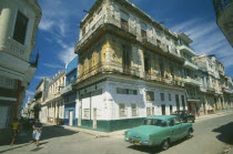 Street corner with traditional buildings with overhanging balconies.  Turquoise car passing in the foreground.  Automobile