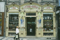 Shop front with mosaic tiling at each side of the doorway and display of dried meat  port and other food items in the windows.  Customers inside and woman walking past. Porto