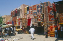 The souk with carpets displayed from walls and awnings of shopfronts opposite street vendors.Market Marrakech Marrakesh