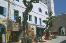 Square with white painted buildings with blue shutters.  Shop with display of Moroccan lanterns and carpets beside cafe with striped awning and outside tables