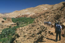 Tourist group walking along narrow  rocky path towards flat roofed housing set into steep hillside with fertile valley gorge on left.