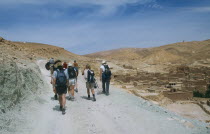 Tourist group following man on horseback along steep stony mountain path in barren landscape with rural buildings on right.