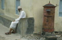 Elderly Muslim man sitting at roadside beside post box. Unguja