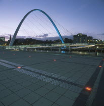 The new Millennium Footbridge illuminated at dusk with city buildings silhouetted against purple evening sky behind.