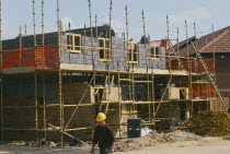 Scaffolding surrounding half completed brick house with man wearing a yellow hard hat in the foreground