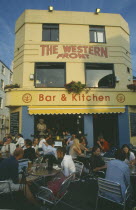 City bar and restaurant with open front and people sitting at crowded outside tables.