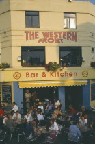 City bar and restaurant with open front and people sitting at crowded outside tables.
