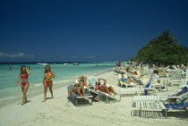 Female tourists beach sunbahing on sun loungers by the water with two women in swimsuits walking past