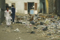 School children walking past rubbish dump with chickens and dogs scavenging in the City of the Dead