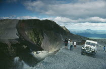 Jeep and visitors parked beside crater rim of volcano.
