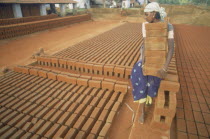 Barefooted young woman carrying large stack of bricks at brick works.