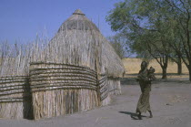 Woman and child in Shilluk village walking past thatched hut and enclosure.