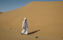 Man in white robe and turban passing huge sand dunes encroaching on village and agricultural land and cutting off road to El Caeid.