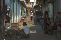 View down a side street with people sat on ground and a woman approaching wearing conical hat.