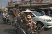 People riding a Cyclo down a busy street with a car beside them