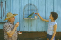 Elderly man and young girl feeding fruit to caged parrot.
