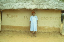 Traditional healer standing outside thatched hut with  Jane is a doctor  written across the wall.