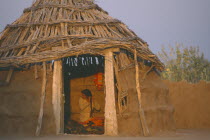 Bhikodai.  Early morning light on thatched mud hut with woman plaiting her hair framed in doorway.rural building