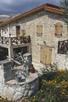 Stone house with overhanging tiled roof and wooden window shutters and door with well in courtyard in the foreground.