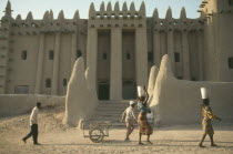 Exterior of mud brick mosque built in traditional style with woman walking past carrying buckets on their heads.