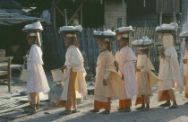 Buddhist nuns wearing white and orange robes carrying trays of wrapped objects on their heads.Burma Myanmar