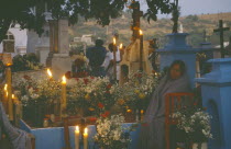 Woman keeping vigil beside grave decorated with candles and flowers during Night of the DeadDay of the Dead