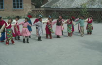 Line of children at kindergarten with linked arms and heads inclined sideways wearing traditional dress.