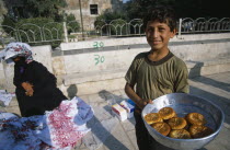 Young boy selling sweetmeats in the market  woman in traditional black dress and veil sitting on pavement behind.