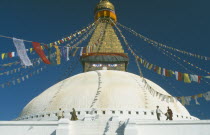Detail of white dome with visitors circling it clockwise and gilded spire hung with prayer flags and painted eyes at its base.