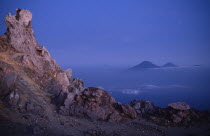 View from the rocky summit of the volcano with peaks in the distance above misty blue blanket of cloud