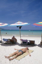 Sun bather lying on the sand in the foreground with row of umbrellas and deck chairs behind and the sea beyond