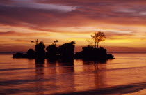Rocky outcrop with trees just off shore silhouetted against dramatic sunset