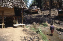 Young Karen refugee man walking in shallow stream toward wood and cane houses built on stilts