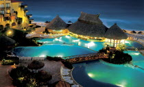 A swimming pool surrounded by sun loungers  wooden thatched huts and bridges lit up at night. Hotel right next to beach.
