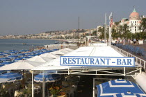View over Restaurant and bar sign along the pebbly beach  Tables with parasols under canopy  blue chairs on promenade.