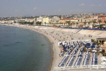 View along the pebbly beach  rows of sun loungers  people swimming and sun bathing. Palm trees line the sea front with shops and buildings.