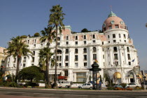 Central reservation with trees and plants.Cars parked in front of pink and white building  many country s Flags flying.