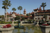 Flagler College  Court yard in the foreground with a water fountain  flowers in stone urns  palm trees and the red and white brick building with towers in the background.