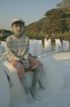 Portrait of a young boy worker sat on bags of cement covered in dust  wearring a baseball cap. Banks of the Irrawaddy Myanmar