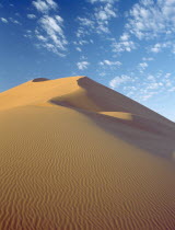 Sand dune with dramatic clouds scattered in a blue sky above seen in early morning light