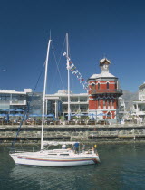 Victoria and Alfred Waterfront. View across water with a yacht sailing past towards a Victorian Gothic style Clock Tower with people sat under parasols along the waterside.Built in 1882