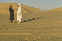 Two men standing in the desert in conversation casting strong shadows across the sand.