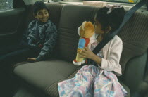 Two Bengali children in back seat of car wearing seat belts.  Young girl showing teddy bear to boy. Automobile