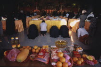 Morning prayers in the main prayer hall. Kneeling worshippers with table in foreground spread with offerings of fruit  flowers and candles.Monastery of Supreme Bliss Center Centre Colored Coloured